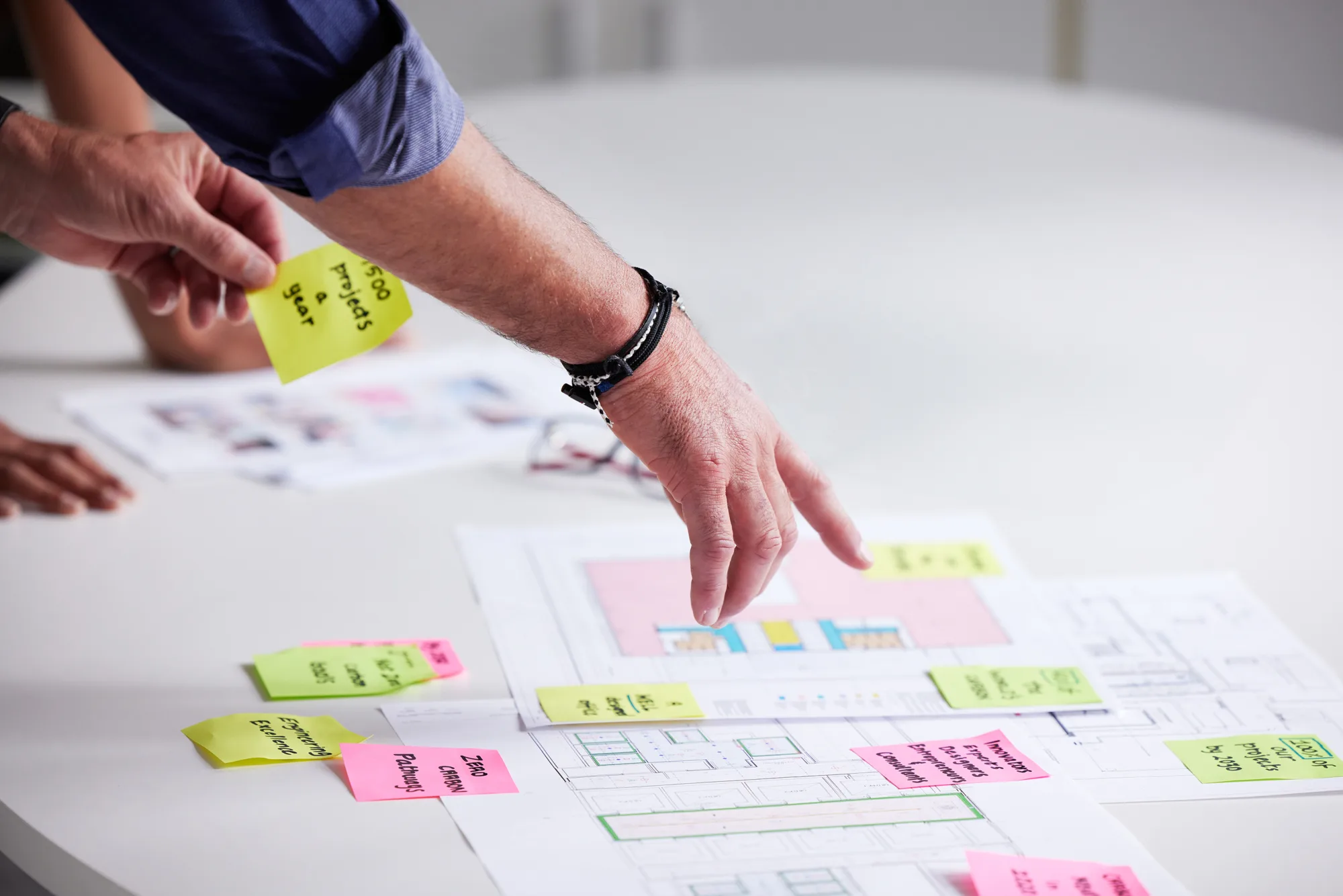 A person’s hand reaches over a table covered with architectural drawings and scattered sticky notes in various colors, while another hand rests nearby at the edge of the table.