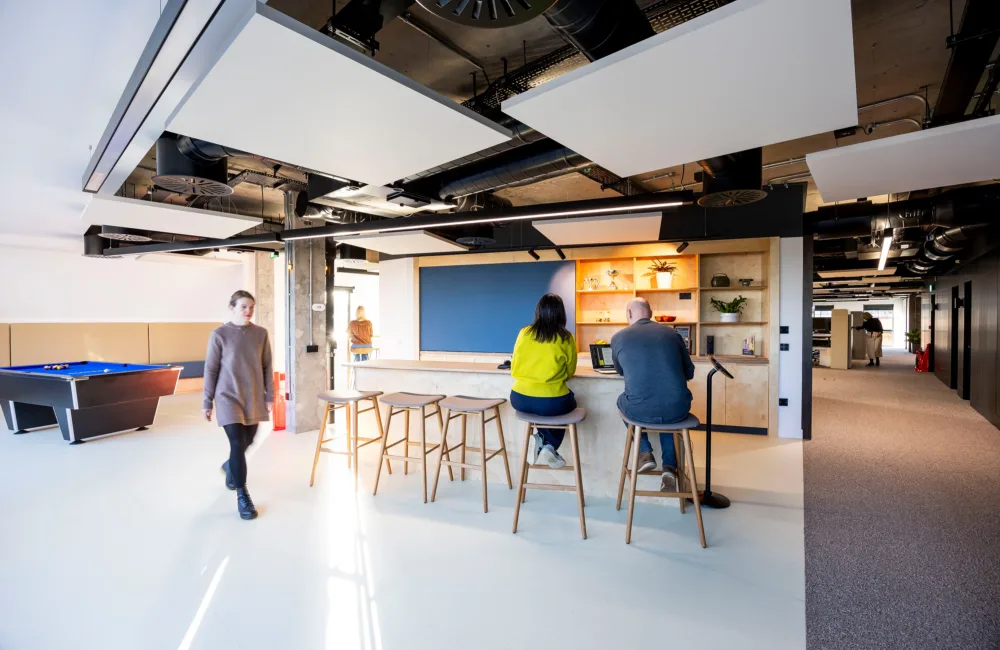 Two people sat on high stools at a countertop in an open plan space with a blue felt pool table and a person walking past towards a long corridor.
