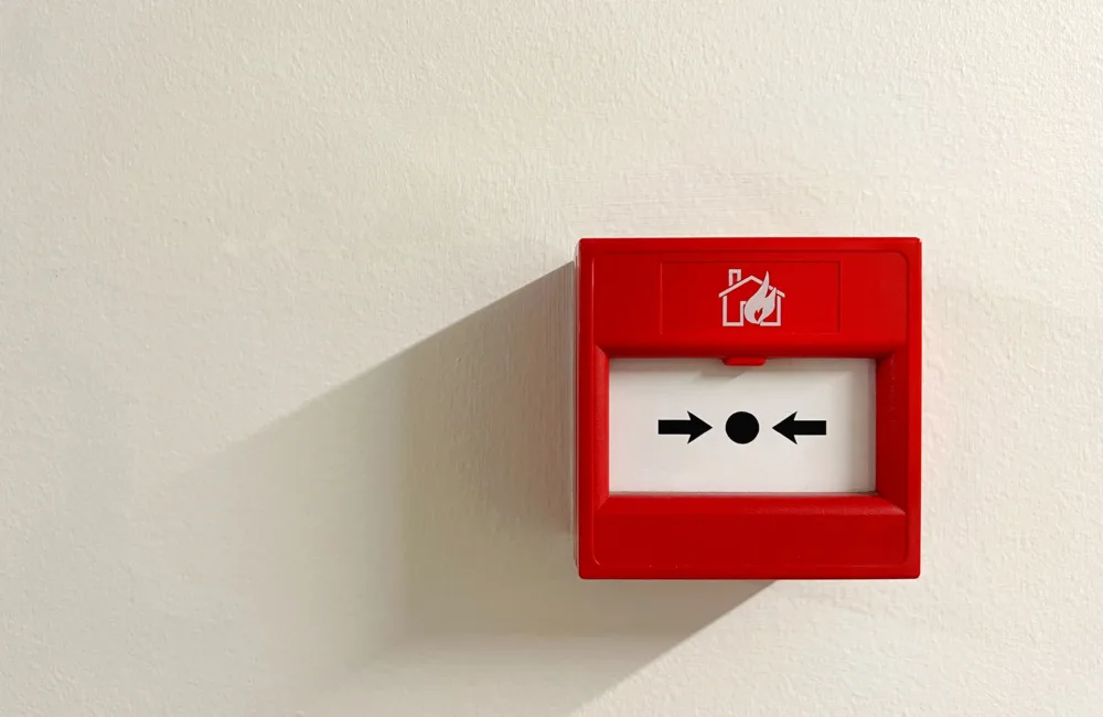 A red fire alarm call point mounted on a white wall. It features a white square with a black dot and two black arrows pointing inward, and an icon of a house with flames above the square.
