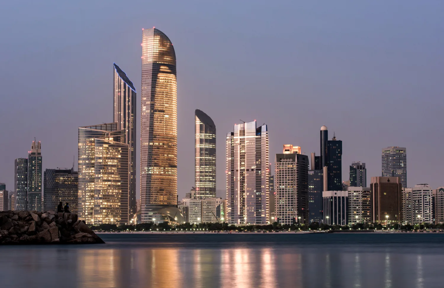 A waterfront city skyline at dusk featuring illuminated high-rise buildings with varied shapes, reflected in calm water, with a rocky shoreline in the foreground and a clear evening sky above.