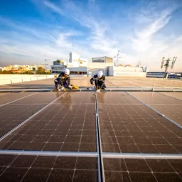 Two workers in safety gear clean solar panels on a rooftop under a blue sky with scattered clouds. Background includes buildings, antennas, and industrial equipment.