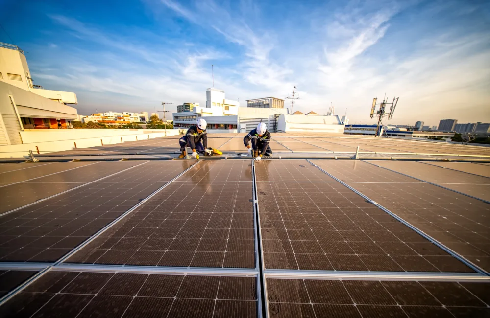Two workers in safety gear clean solar panels on a rooftop under a blue sky with scattered clouds. Background includes buildings, antennas, and industrial equipment.