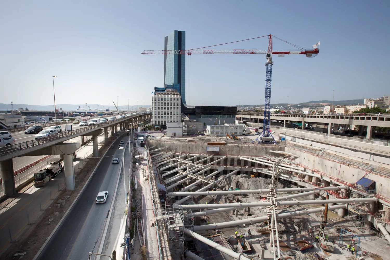Large urban construction site with deep excavation and structural supports; tall building and crane in background; elevated highways with moving vehicles on both sides.