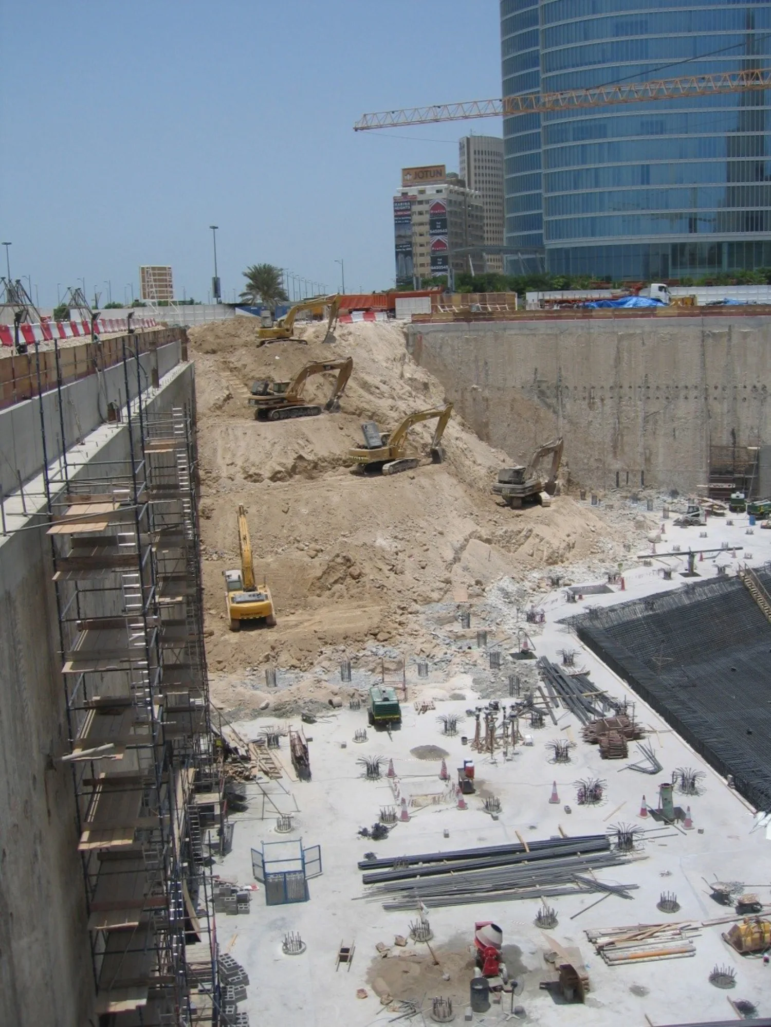 Large construction site with multiple excavators working on a dirt pile; surrounded by tall buildings including a glass skyscraper; scaffolding and construction materials visible across the site.