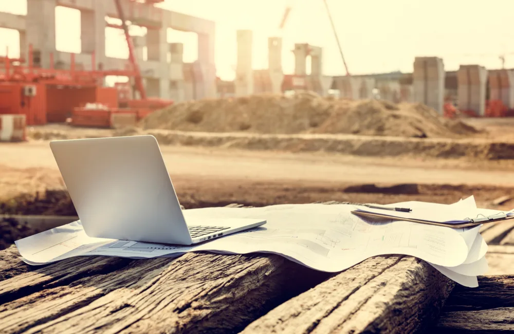 Construction site with a partially built structure in the background; foreground features a wooden surface with an open laptop and large sheets of paper resembling blueprints; heavy machinery and materials visible.