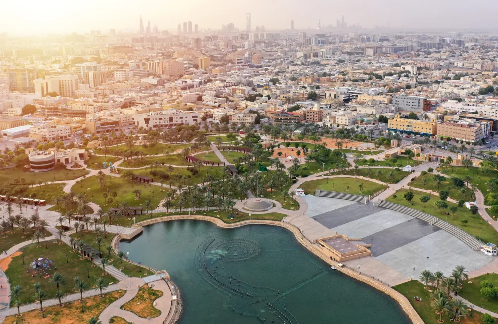 Aerial view of a cityscape with tall buildings in the background and a large park in the foreground, featuring green lawns, pathways, palm trees, a pond with fountains, and recreational zones.