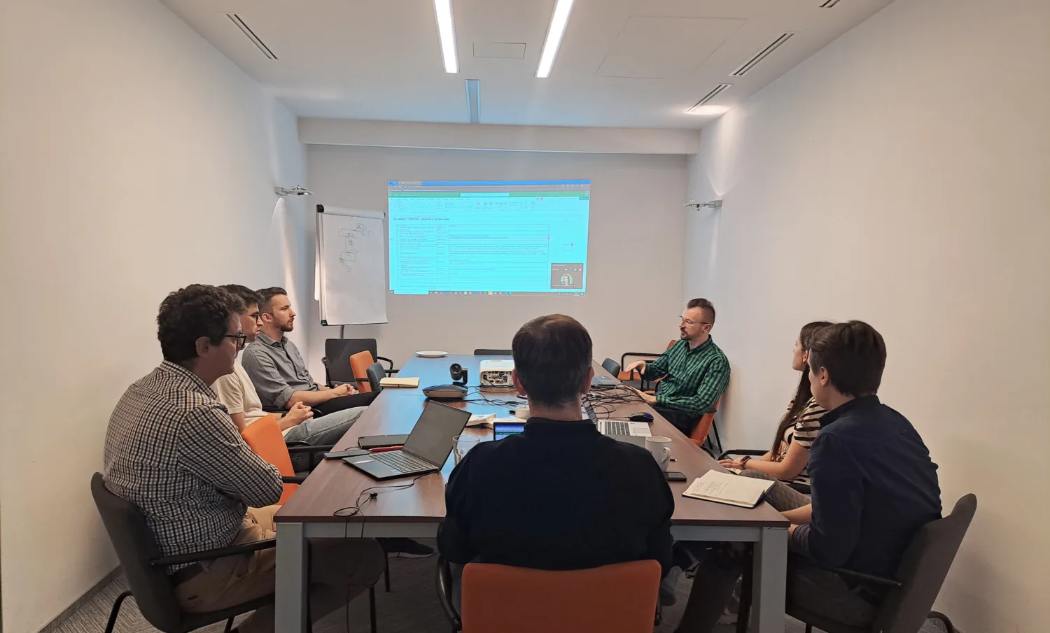 Speaker standing in meeting room presenting with projection of presentation on wall to small group around a table