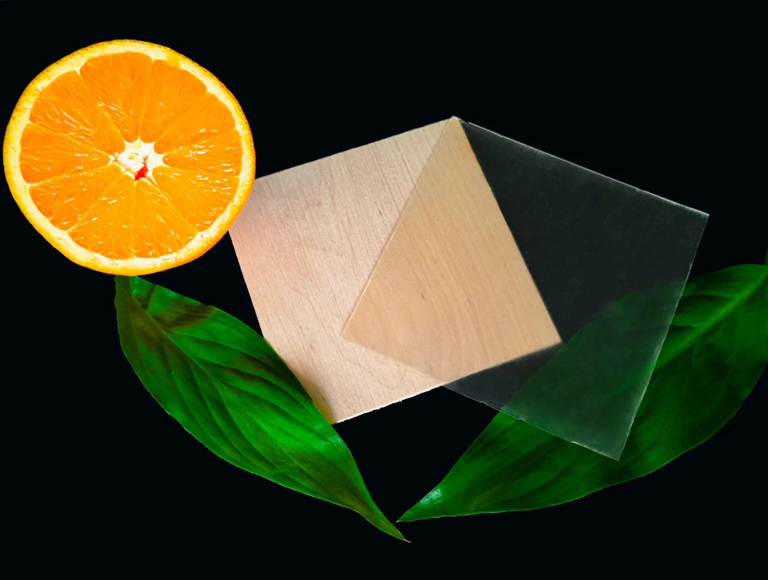 A square sheet of transparent wood on a black surface on top of a wooden square, sliced orange and green leaf