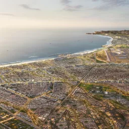Aerial view of a coastal city with dense residential blocks, intersecting roads, and open plots beside a curved shoreline. Buildings extend toward nearby hills, with a harbour, beach, and calm water under a hazy sky.