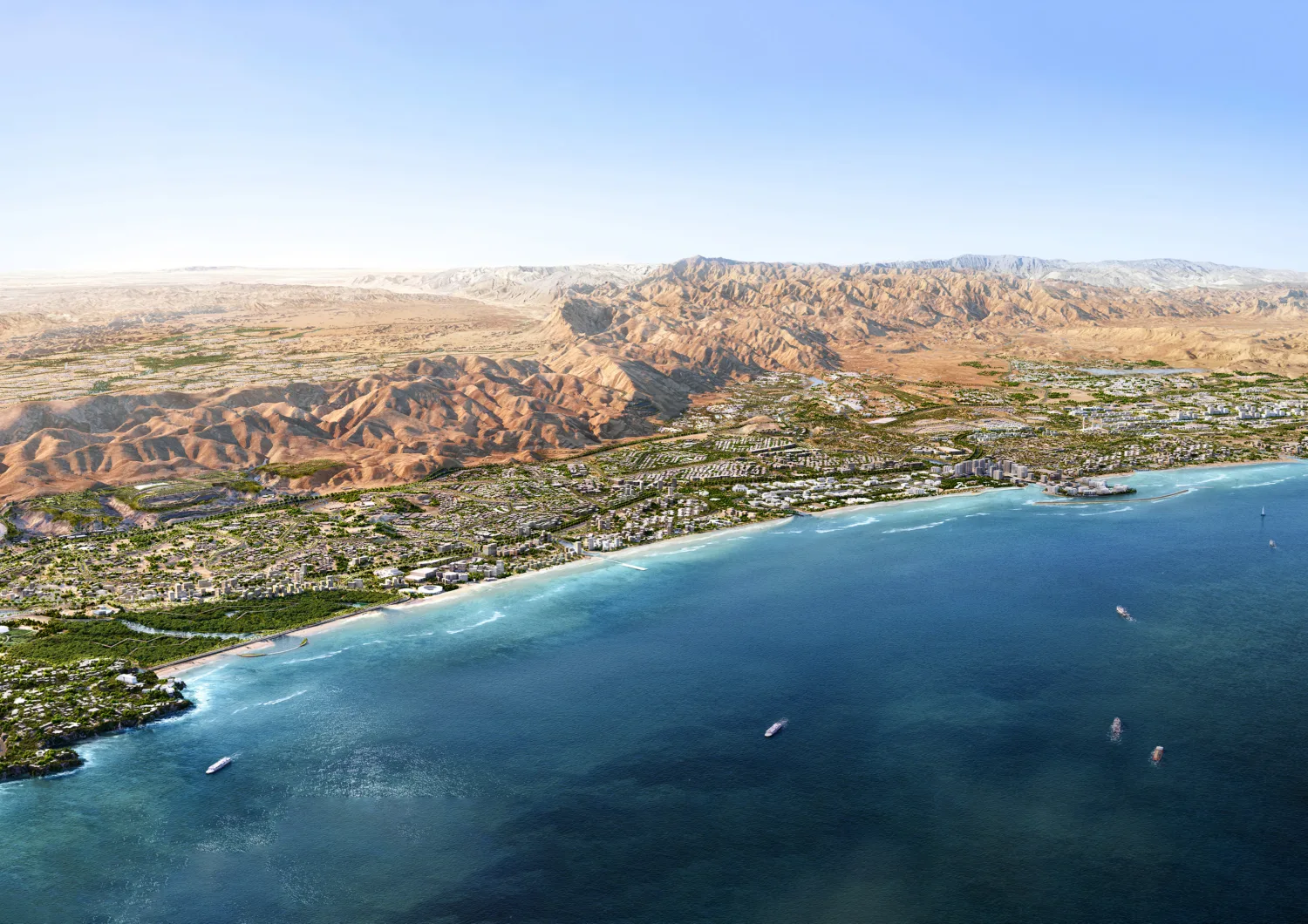 Aerial view of a coastal city with dense low- and mid-rise buildings along a curved shoreline, turquoise water with several boats, sandy beaches, and extensive brown mountains stretching inland under a clear sky.