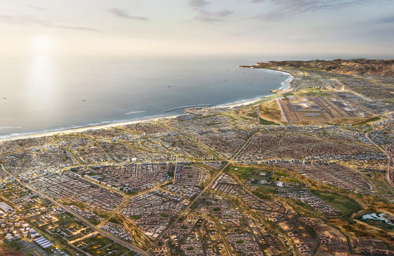 Aerial view of a coastal city with dense residential blocks, intersecting roads, and open plots beside a curved shoreline. Buildings extend toward nearby hills, with a harbour, beach, and calm water under a hazy sky.