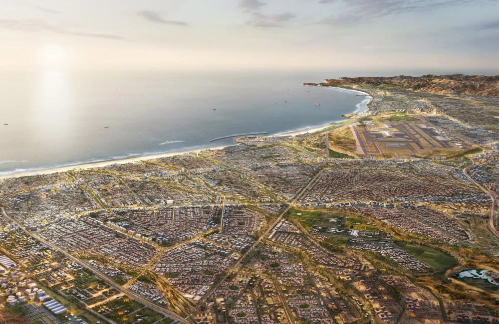 Aerial view of a coastal city with dense residential blocks, intersecting roads, and open plots beside a curved shoreline. Buildings extend toward nearby hills, with a harbour, beach, and calm water under a hazy sky.