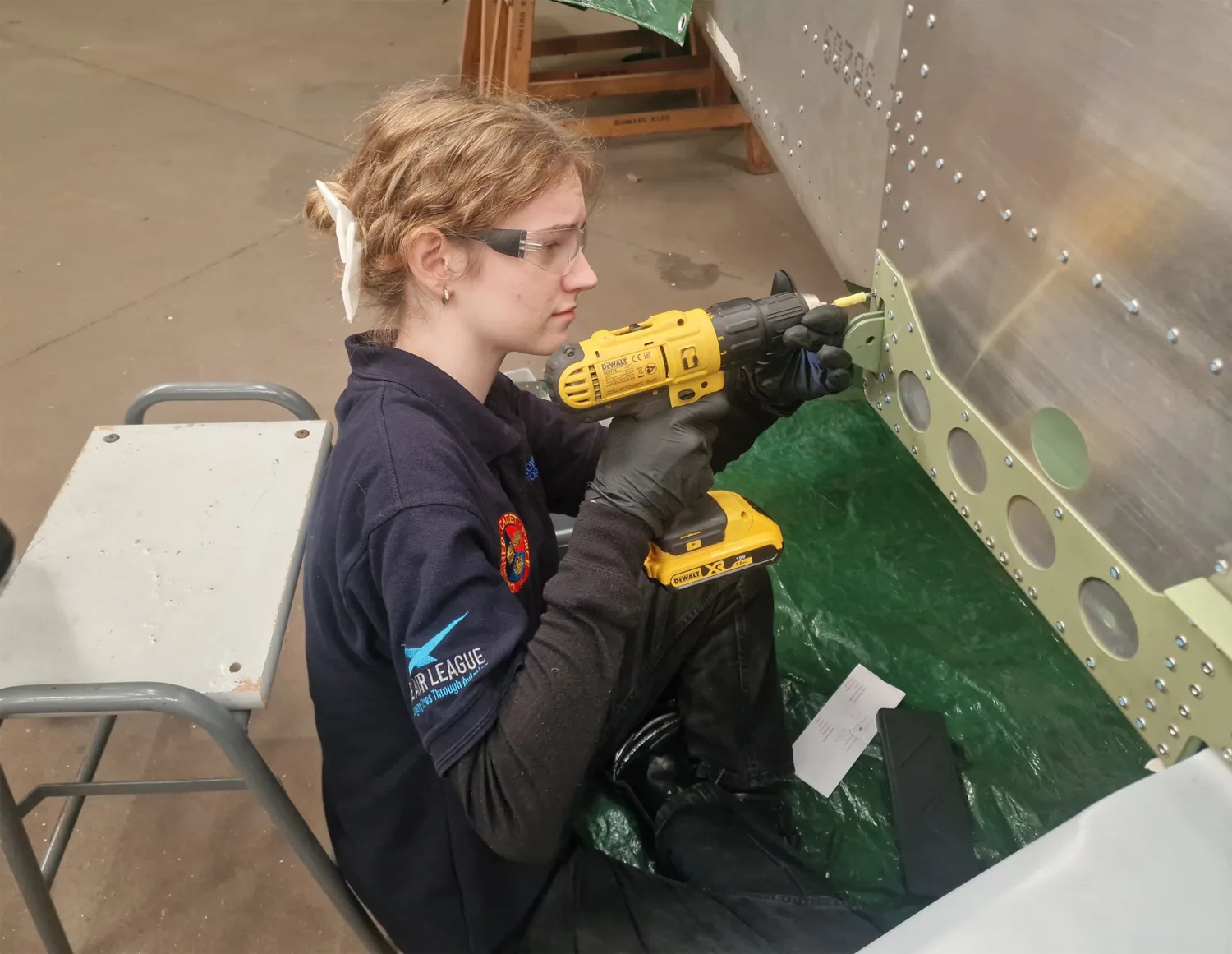 Person seated on a low stool uses a yellow cordless drill to fasten a green metal panel to an aluminium aircraft fuselage. Wearing gloves and workwear, the scene shows a workshop floor, tools, rivets, and protective sheeting nearby.