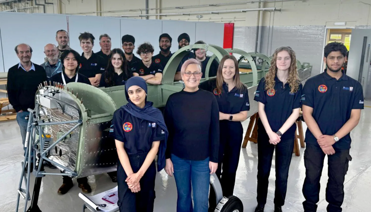 Group of people wearing dark polo shirts standing inside an industrial workshop beside a partially assembled aircraft fuselage. The space includes metal structures, tools, workbenches and overhead lighting.
