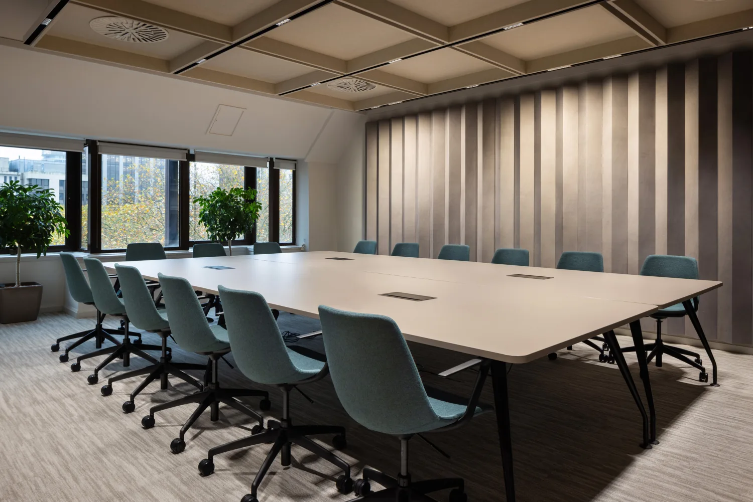 Modern conference room with a large rectangular table surrounded by 12 teal office chairs on wheels, striped wall, three large windows with potted plants, and beige carpeted floor.