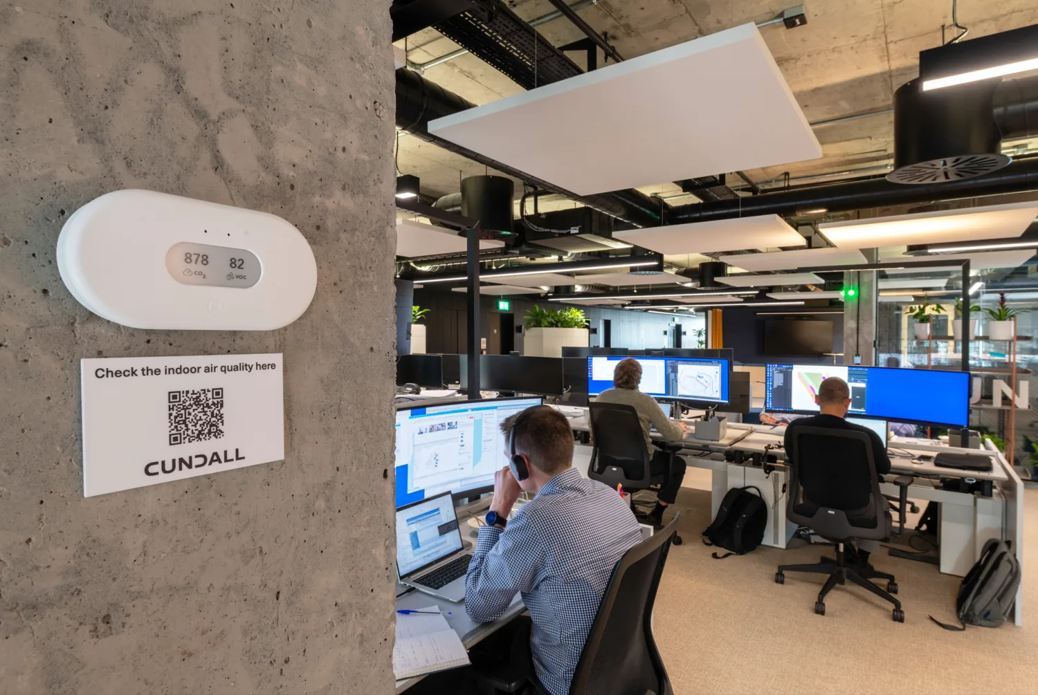 Modern office with three people at desks using monitors. A device on a concrete pillar displays “878 ppm CO₂” and “82% humidity.” Below is a sign reading “Check the indoor air quality here” with a QR code and “CUNDALL.”