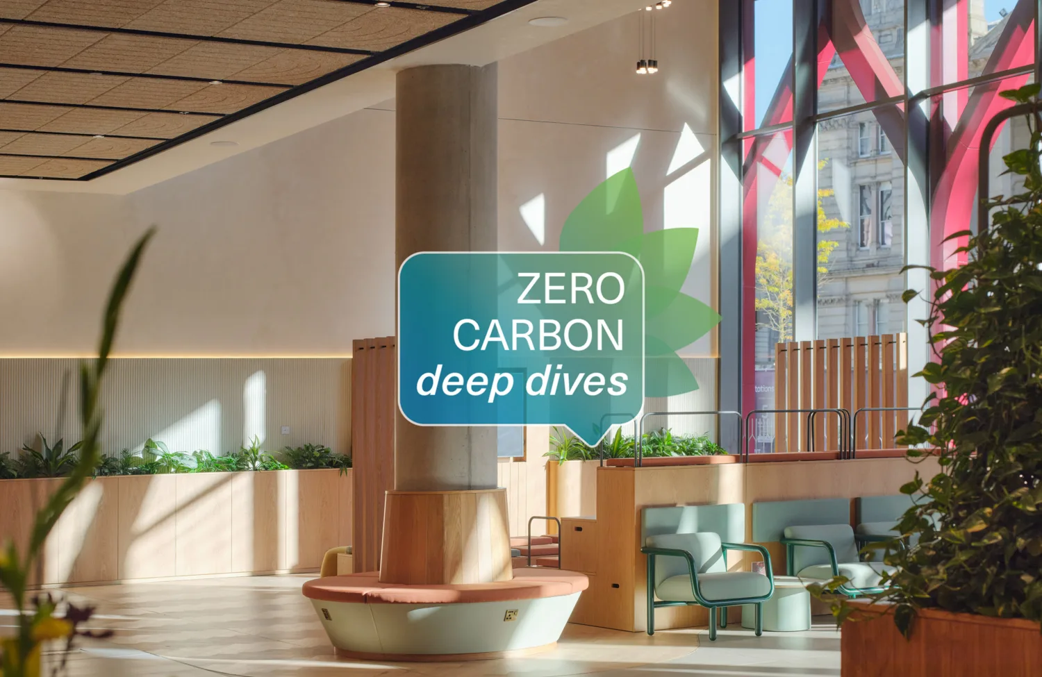 Bright, modern interior atrium with timber seating, planters, and green chairs around a central column. Large windows reveal an urban street outside. Overlaid graphic reads ‘ZERO CARBON deep dives’.
