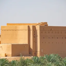 Large rectangular adobe building with smooth tan walls featuring rows of triangular and rectangular ventilation openings. Smaller tan structures are adjacent. Palm trees in the foreground and arid terrain in the background under a clear sky.