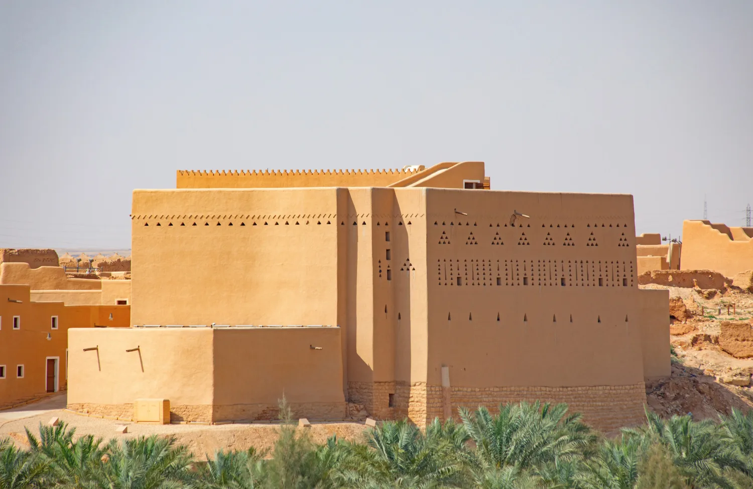 Large rectangular adobe building with smooth tan walls featuring rows of triangular and rectangular ventilation openings. Smaller tan structures are adjacent. Palm trees in the foreground and arid terrain in the background under a clear sky.