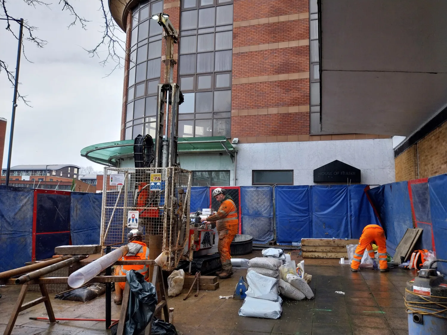 Construction site with workers in orange safety gear operating machinery near a tall drilling rig, enclosed by blue and red barriers. A brick building in the background displays the sign "House of Fraser."