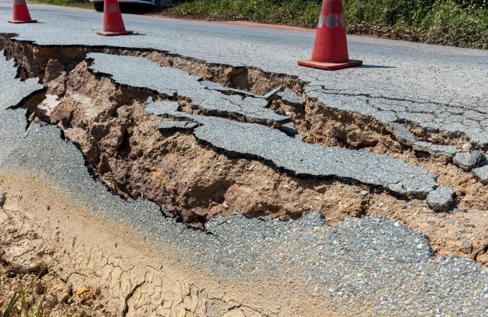 A severely cracked and eroded asphalt road with large fissures and exposed soil, spanning the full width. Three orange traffic cones are placed near the damaged area to signal caution.