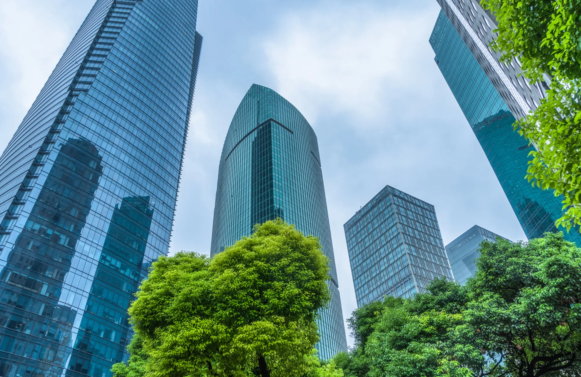 A group of modern glass-clad high-rise buildings viewed from below, with reflective facades under a cloudy sky, surrounded by dense green trees and foliage in the foreground.