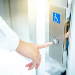Close-up of a hand pressing an elevator call button on a stainless steel panel. A blue wheelchair accessibility symbol is displayed above the round button, next to an open lift door.