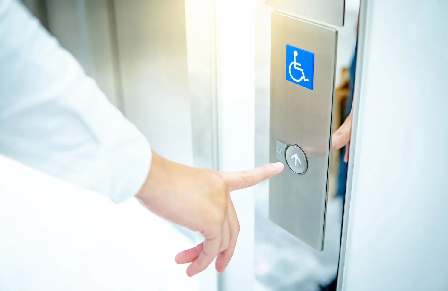 Close-up of a hand pressing an elevator call button on a stainless steel panel. A blue wheelchair accessibility symbol is displayed above the round button, next to an open lift door.
