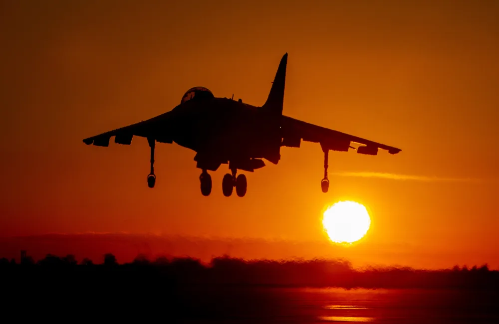 A military jet with landing gear extended is silhouetted against an orange sunset, with the sun low on the horizon and light reflecting off the ground.