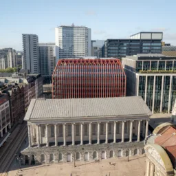 Aerial view of a city centre with a neoclassical building featuring a columned façade in the foreground, behind a modern rectangular building with red lattice cladding, surrounded by glass offices and taller residential blocks.