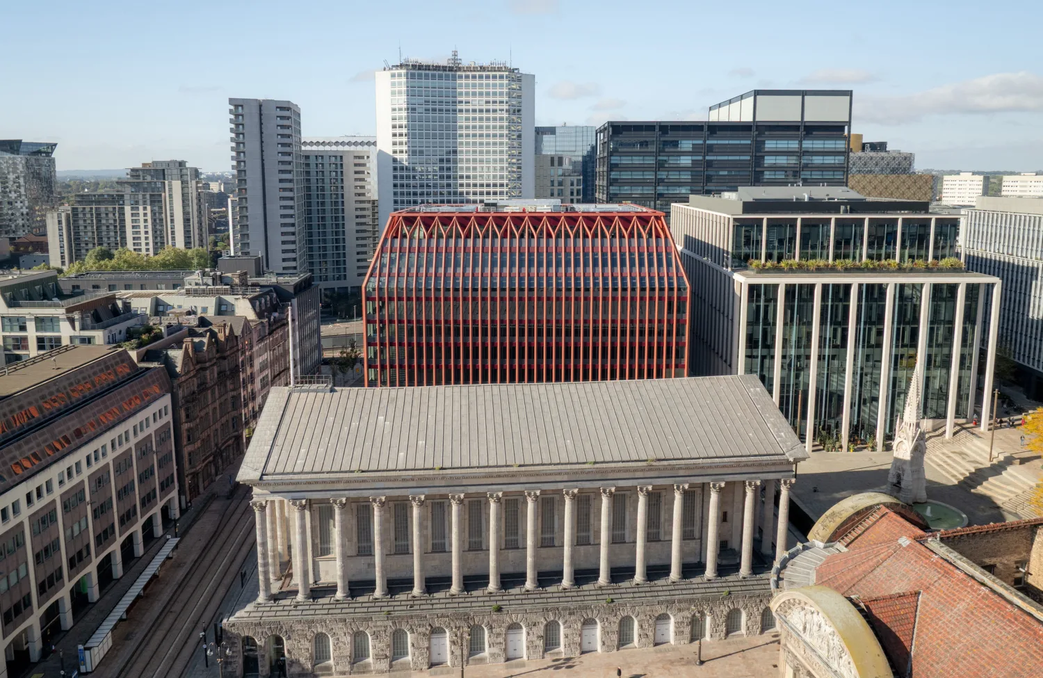 Aerial view of a city centre with a neoclassical building featuring a columned façade in the foreground, behind a modern rectangular building with red lattice cladding, surrounded by glass offices and taller residential blocks.