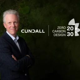 A person in a dark checked blazer stands with folded arms in front of a green foliage wall. The Cundall logo appears beside them, along with the text “Zero Carbon Design 2030” and a stylized leaf graphic.