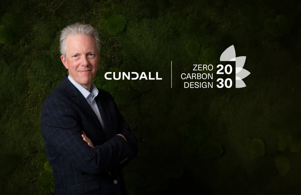 A person in a dark checked blazer stands with folded arms in front of a green foliage wall. The Cundall logo appears beside them, along with the text “Zero Carbon Design 2030” and a stylized leaf graphic.