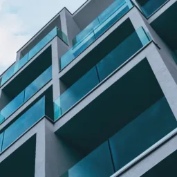 A low-angle view of a modern multi‑storey building with staggered balconies. Each balcony has turquoise-tinted glass railings and smooth grey walls, set against a partly cloudy sky.