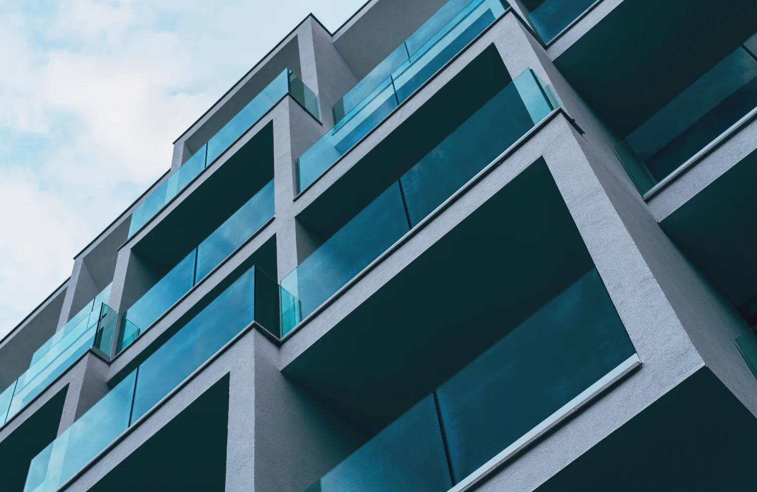 A low-angle view of a modern multi‑storey building with staggered balconies. Each balcony has turquoise-tinted glass railings and smooth grey walls, set against a partly cloudy sky.