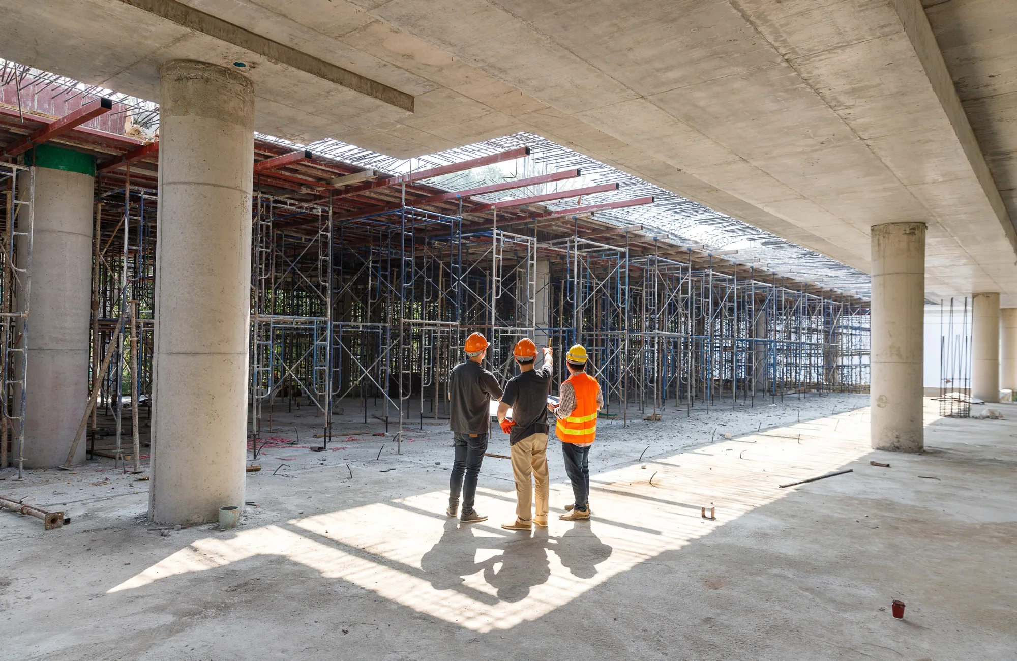 Three construction workers in orange safety helmets and vests stand beneath a large concrete structure with scaffolding and support columns. Sunlight filters through ceiling gaps, casting shadows.