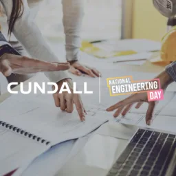 Close-up of three people’s hands pointing at technical drawings on a desk with an open spiral notebook and a laptop. A yellow hard hat and documents are in the background. Visible text includes “CUNDALL” and “National Engineering Day.
