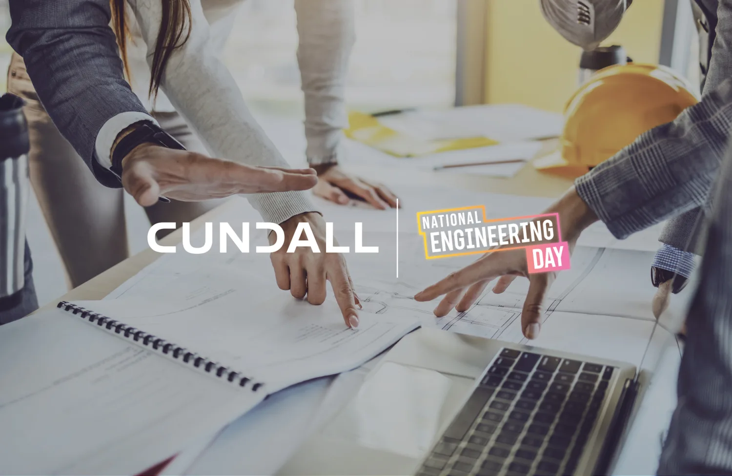 Close-up of three people’s hands pointing at technical drawings on a desk with an open spiral notebook and a laptop. A yellow hard hat and documents are in the background. Visible text includes “CUNDALL” and “National Engineering Day.