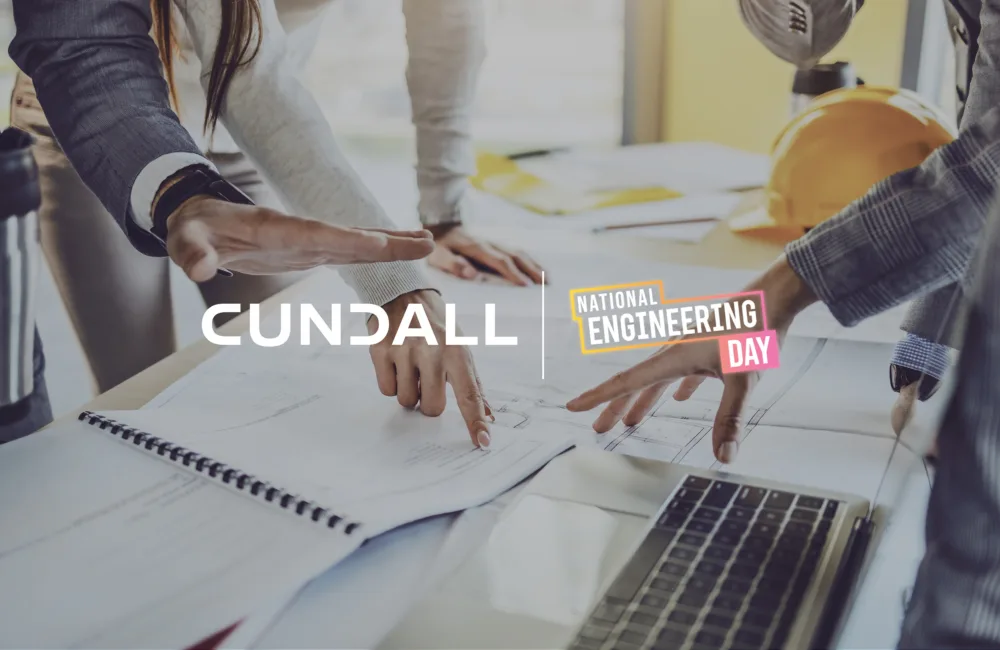Close-up of three people’s hands pointing at technical drawings on a desk with an open spiral notebook and a laptop. A yellow hard hat and documents are in the background. Visible text includes “CUNDALL” and “National Engineering Day.