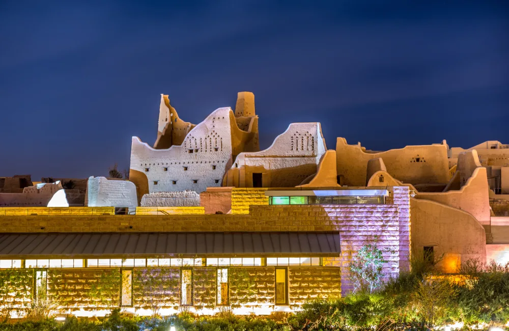 Historic mud-brick ruins with tall, irregular walls and patterned openings lit at night, with a modern flat-roofed building featuring glass panels and stone walls in the foreground under a dark blue sky.