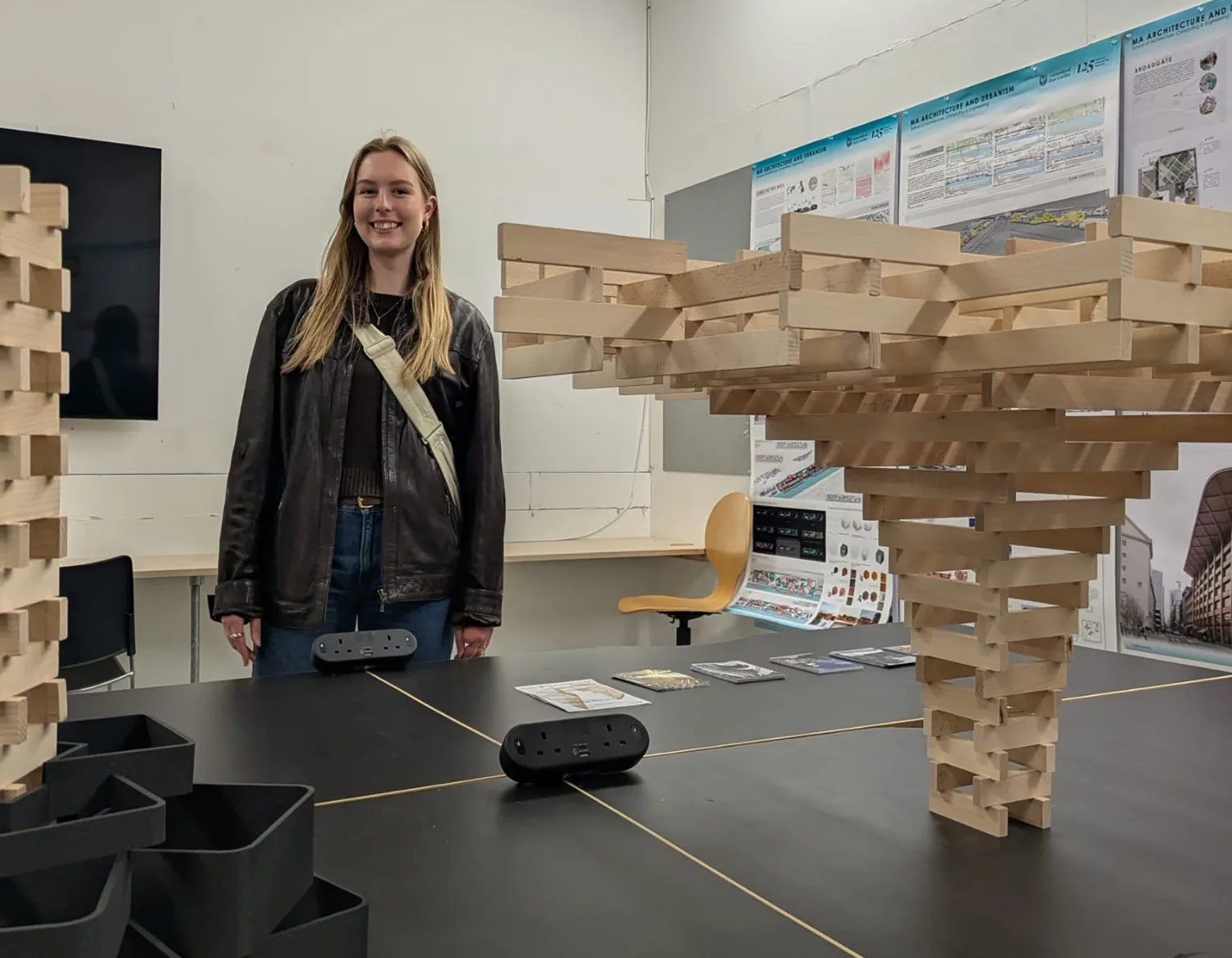 A person stands behind a table displaying wooden block structures resembling architectural models; background includes posters with diagrams and text, a yellow chair, and a wall-mounted monitor.