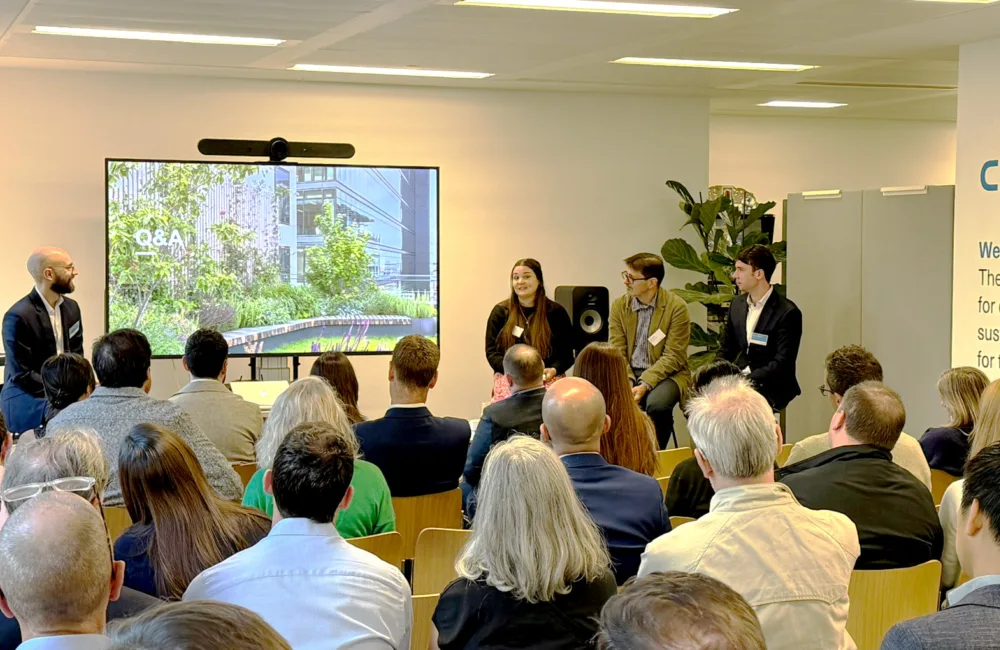 A group of people seated in a conference room face a panel of four speakers. A screen displays “Q&A” with greenery and buildings. A wall reads: “CUNDALL...sustainable solutions for the planet.”