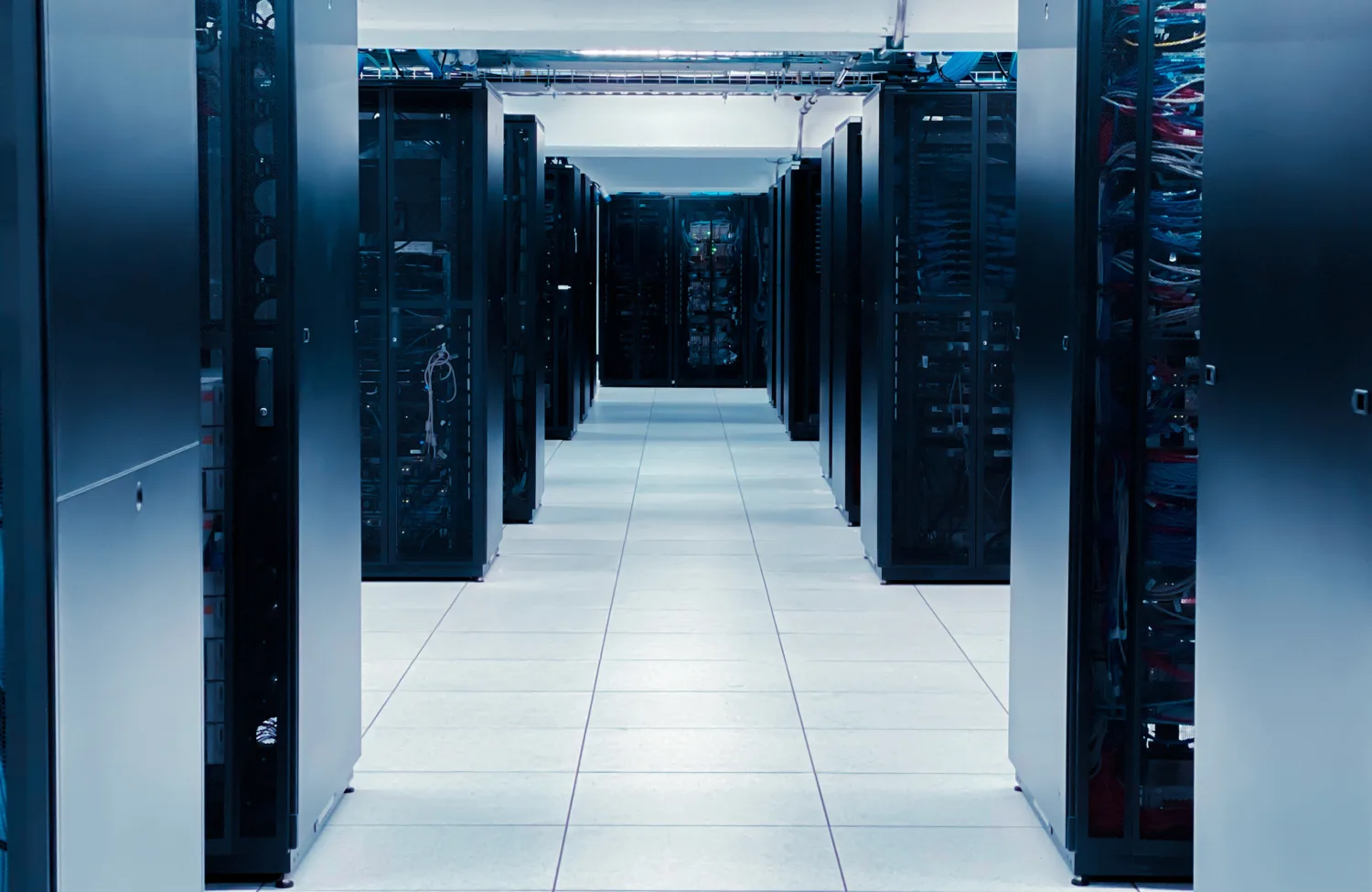 Interior of a data centre with multiple rows of tall black server racks on both sides of a wide aisle, white tiled floor, and overhead cable trays with visible wiring.