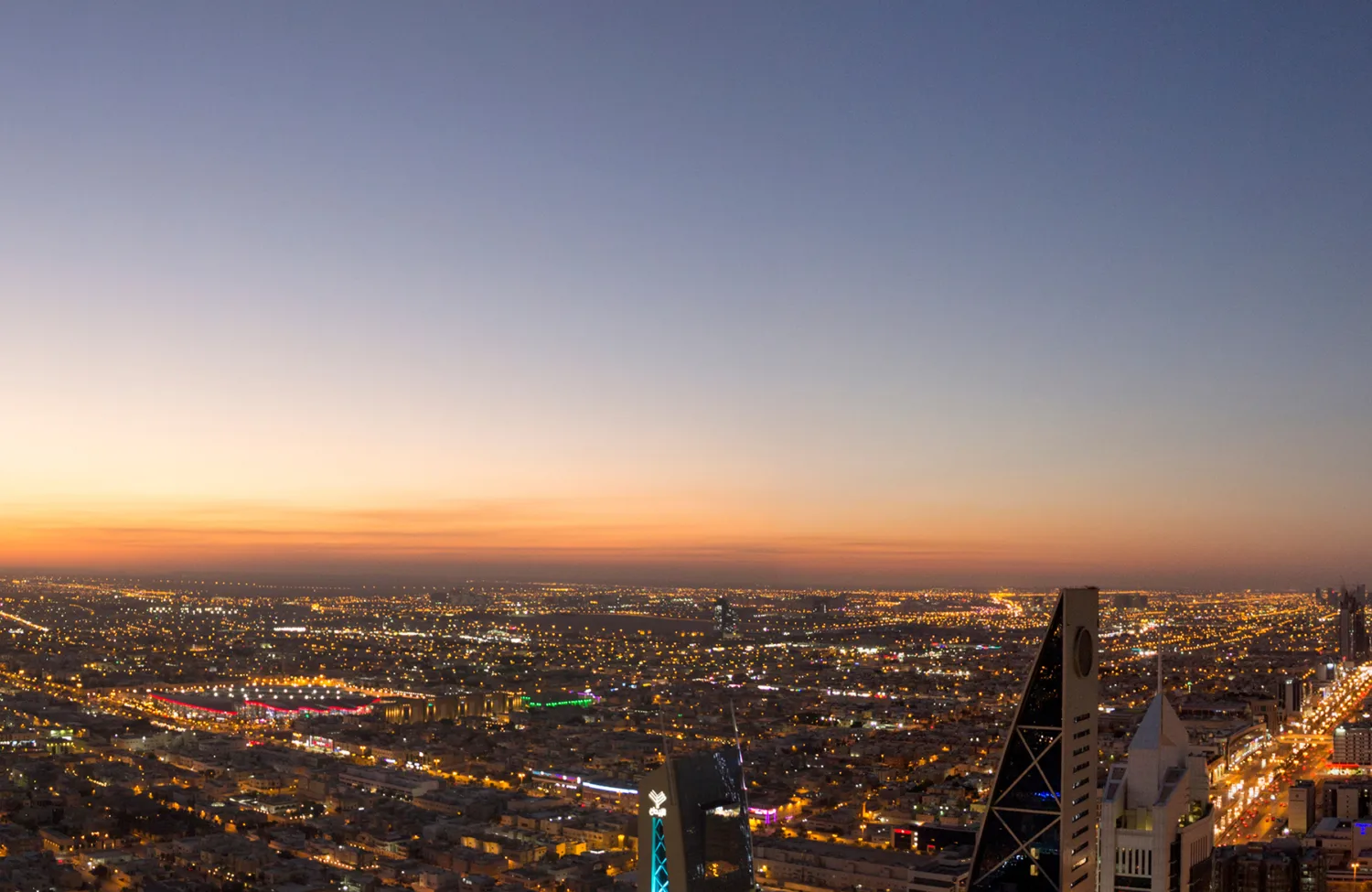 Wide panoramic view of a city at dusk, showing dense illuminated streets and high‑rise buildings. A cluster of tall towers lines a brightly lit avenue on the right, with a gradient sky transitioning from orange near the horizon to blue above.