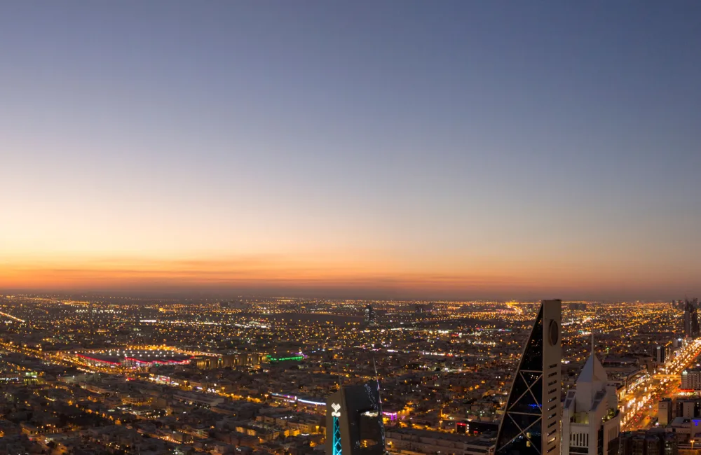 Wide panoramic view of a city at dusk, showing dense illuminated streets and high‑rise buildings. A cluster of tall towers lines a brightly lit avenue on the right, with a gradient sky transitioning from orange near the horizon to blue above.