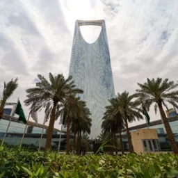 A tall, modern skyscraper with a distinctive open top rises behind rows of palm trees and green landscaping under a cloudy sky, with nearby glass buildings and flagpoles visible.