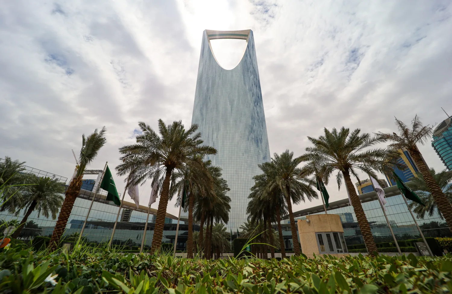 A tall, modern skyscraper with a distinctive open top rises behind rows of palm trees and green landscaping under a cloudy sky, with nearby glass buildings and flagpoles visible.