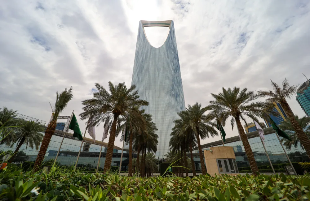 A tall, modern skyscraper with a distinctive open top rises behind rows of palm trees and green landscaping under a cloudy sky, with nearby glass buildings and flagpoles visible.