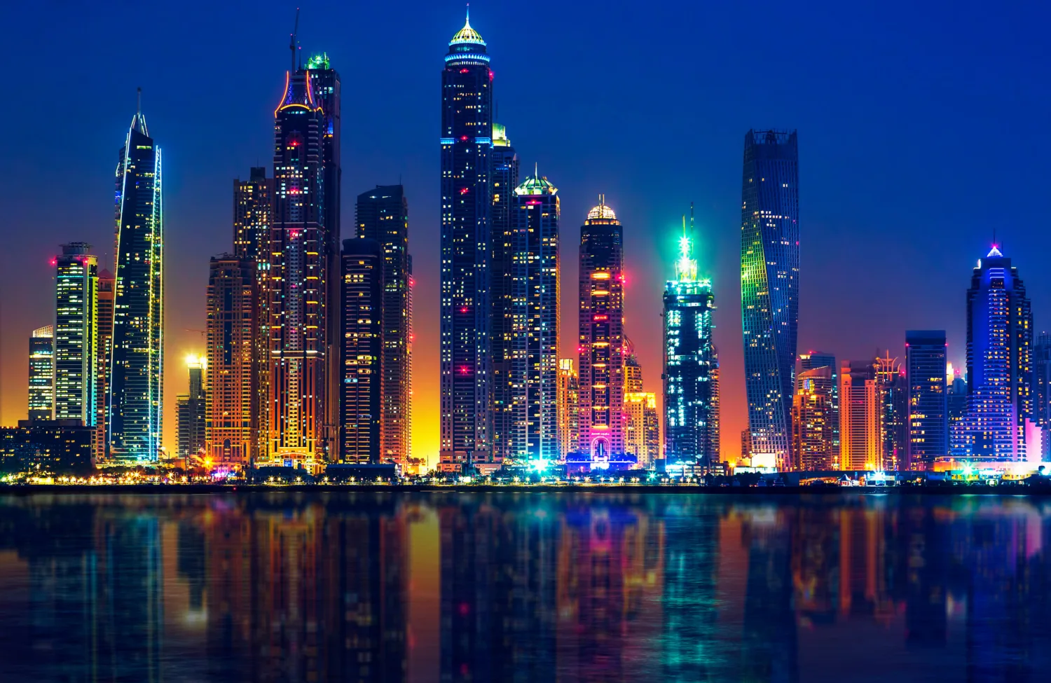 Nighttime city skyline with numerous illuminated high‑rise buildings of varying shapes, including one twisted tower. Bright multi-coloured lights reflect across calm water in the foreground under a dark blue sky.