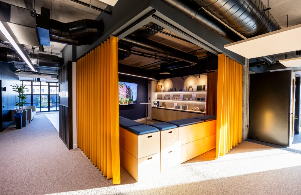 Open-layout office with yellow curtains, wooden benches with blue cushions, wall-mounted TV showing colourful content, shelves with items, exposed ceiling ductwork, and large windows with natural light.